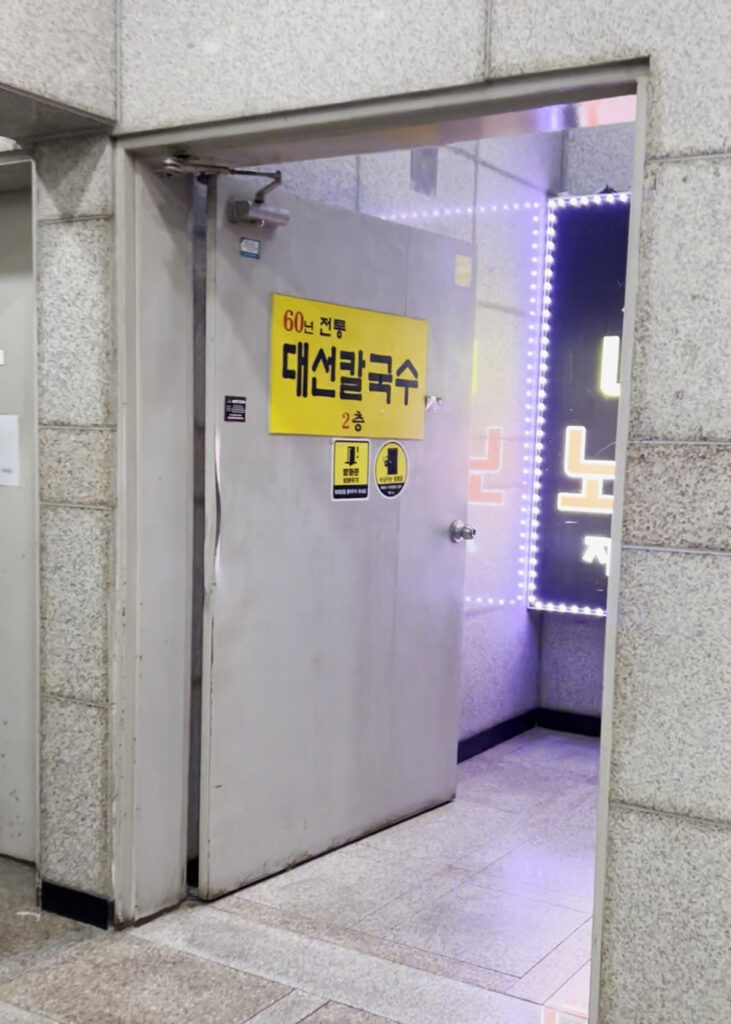 '60년 전통 대선칼국수 2층'이라는 안내문이 붙어있는 가게 입구. The entrance to Daeseon Kalguksu, with a sign that reads '60 years of tradition, Daeseon Kalguksu 2nd floor'.