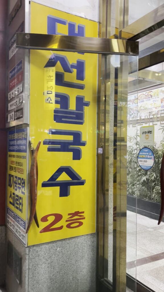 '대전칼국수 2층'이라고 쓰인 대선칼국수의 노란색 복도 간판. The yellow hallway sign for Daeseon Kalguksu in Daejeon, indicating the restaurant is on the 2nd floor.