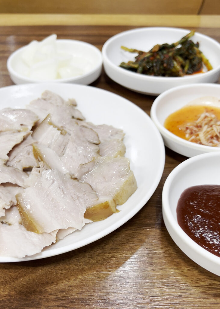 부드럽게 삶아진 돼지고기 수육 한 접시와 함께 나온 김치 및 양념장. A plate of tender boiled pork suyuk served with kimchi and various dipping sauces at Daeseon Kalguksu.