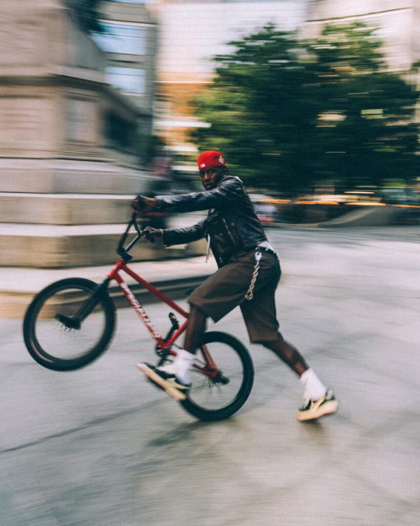 나이젤 실베스터가 BMX 자전거로 윌리 기술을 선보이며 에어 조던 1 로우를 신고 있는 역동적인 모습. A dynamic action shot of Nigel Sylvester doing a wheelie on his BMX bike while wearing the Air Jordan 1 Lows.