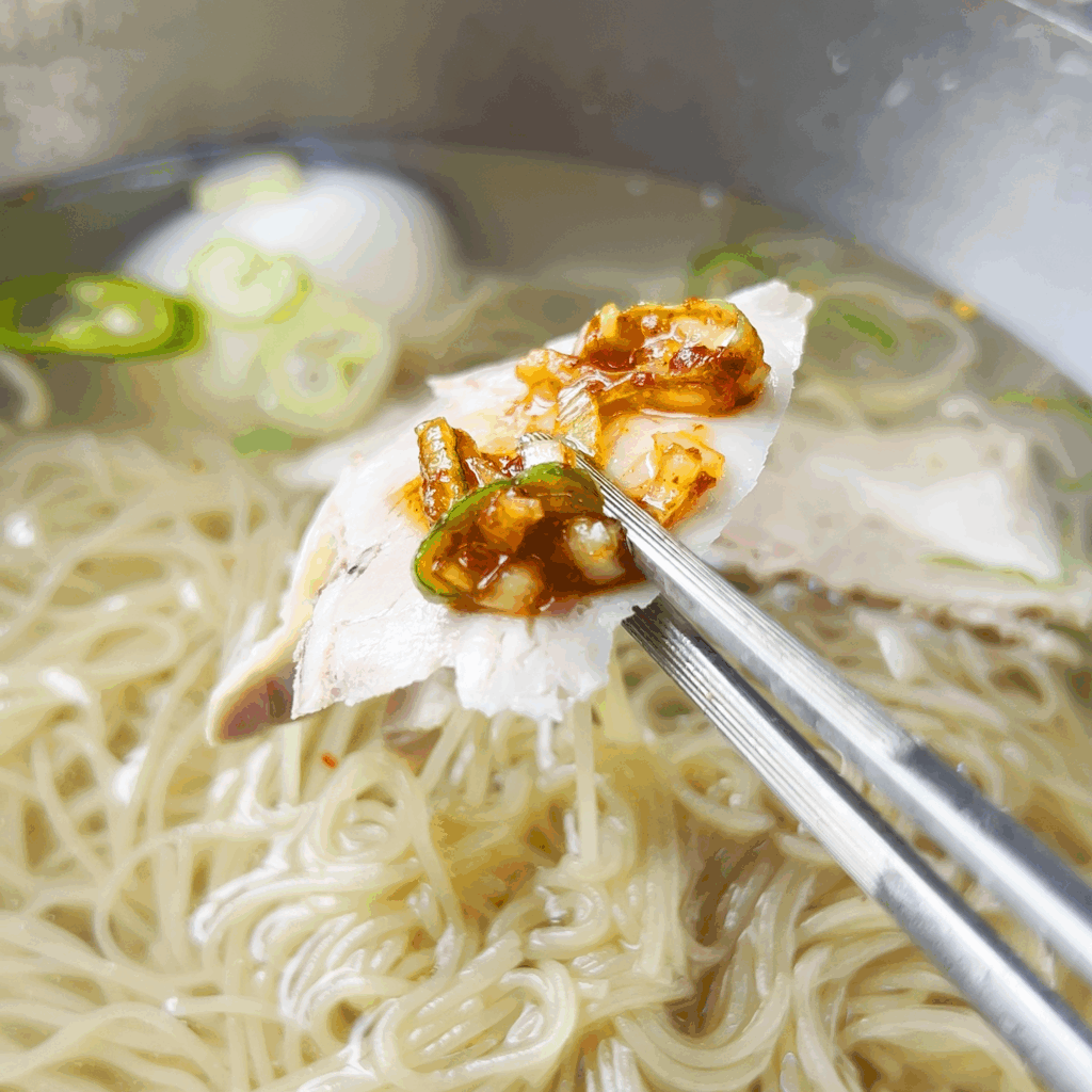 Chopsticks holding a piece of suyuk (boiled pork) covered in Pildong Myeonok's special dipping sauce, hovering over a bowl of Pyeongyang naengmyeon.