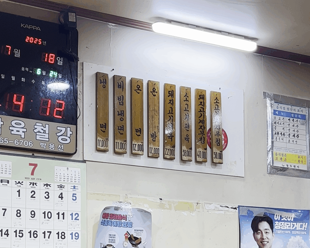 나무판에 세로로 쓰인 평남면옥의 메뉴판. 물냉면, 비빔냉면, 돼지고기편육 등의 메뉴와 가격이 보인다. The wooden menu board at Pyeongnam Myeonok, showing dishes like Mul-naengmyeon, Bibim-naengmyeon, and pork pyeonyuk with their prices.