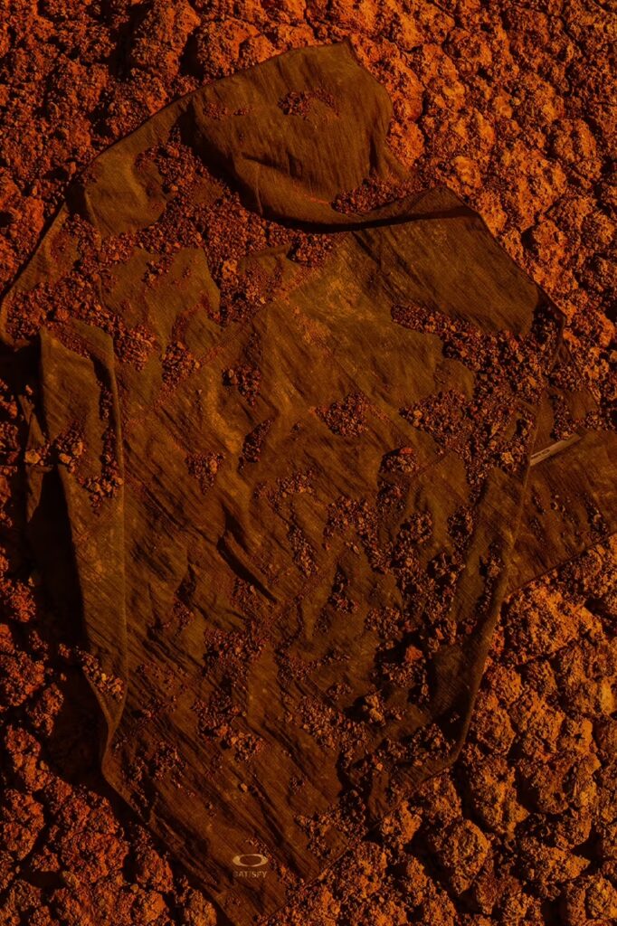 흙먼지 위에 펼쳐진 새티스파이 x 오클리 오라라이트 베이스 레이어. The Satisfy x Oakley Auralite base layer spread out on dusty earth.