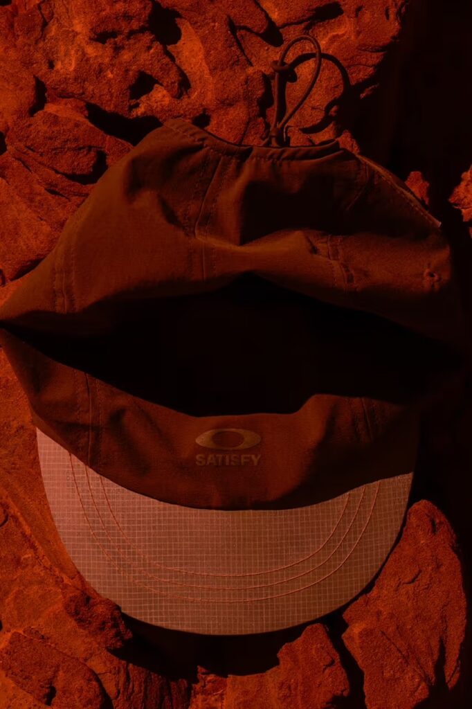붉은 흙 위에 놓인 새티스파이 x 오클리 피스쉘 트레일 캡. The Satisfy x Oakley PeaceShell trail cap placed on a bed of red earth.