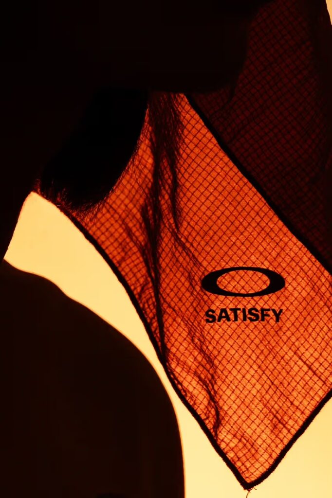 빛에 비춰 격자무늬가 드러나는 립스톱 소재의 새티스파이 x 오클리 반다나. The Satisfy x Oakley ripstop bandana held up to the light, showing the grid pattern of the fabric.