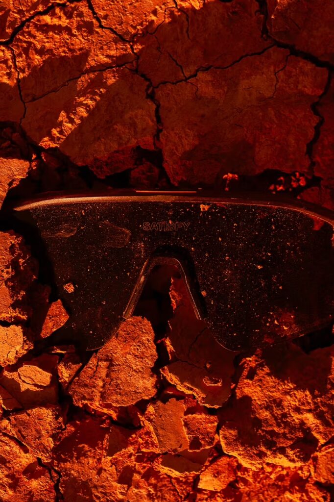 붉은 흙과 돌 위에 놓인 새티스파이 x 오클리 '스턴트 데빌' 선글라스의 정면. A front view of the Satisfy x Oakley 'Stunt Devil' sunglasses resting on red earth and rocks.