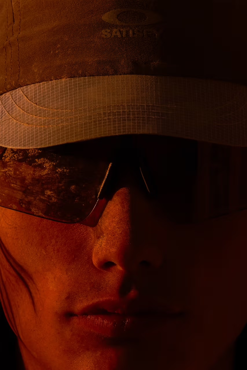 새티스파이 x 오클리 트레일 캡과 '스턴트 데빌' 선글라스를 착용한 모델의 얼굴 클로즈업. A close-up shot of a model wearing the Satisfy x Oakley trail cap and 'Stunt Devil' sunglasses.