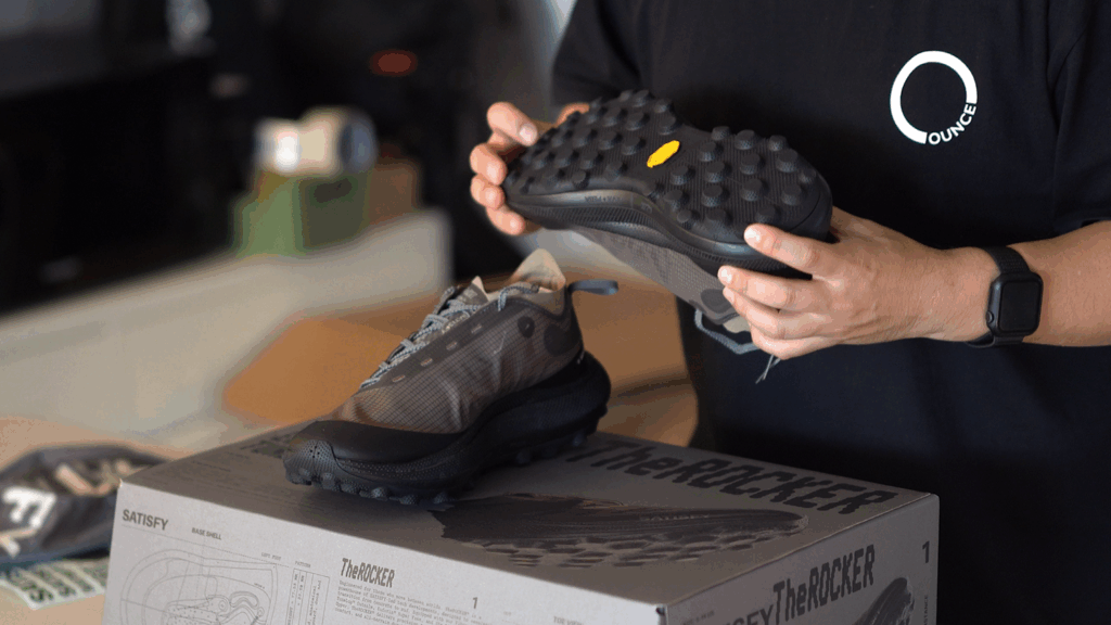 비브람(Vibram) 로고가 보이는 새티스파이 더 락 팔콘의 접지력 좋은 아웃솔(밑창)을 보여주는 모습. A person showcasing the high-traction Vibram outsole of the Satisfy The Rock Falcon shoe.