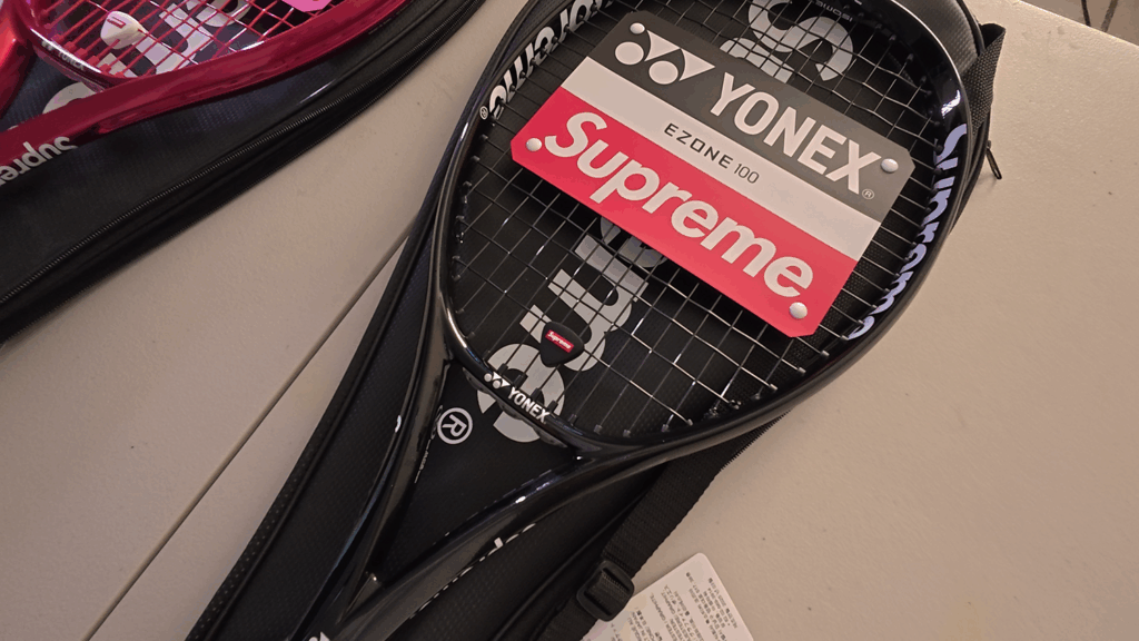 슈프림 로고가 새겨진 라켓 커버 위에 놓인 슈프림 x 요넥스 EZONE 블랙 테니스 라켓. The black Supreme x Yonex EZONE tennis racket resting on its black cover featuring the Supreme logo.