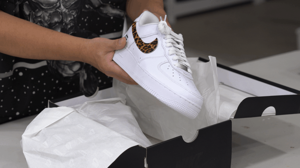 박스에서 슈프림 x 굿이너프 에어 포스 1 화이트 레오파드 스니커즈를 꺼내는 모습. A person unboxing and lifting the white and leopard print Supreme x Goodenough Air Force 1 from its box.