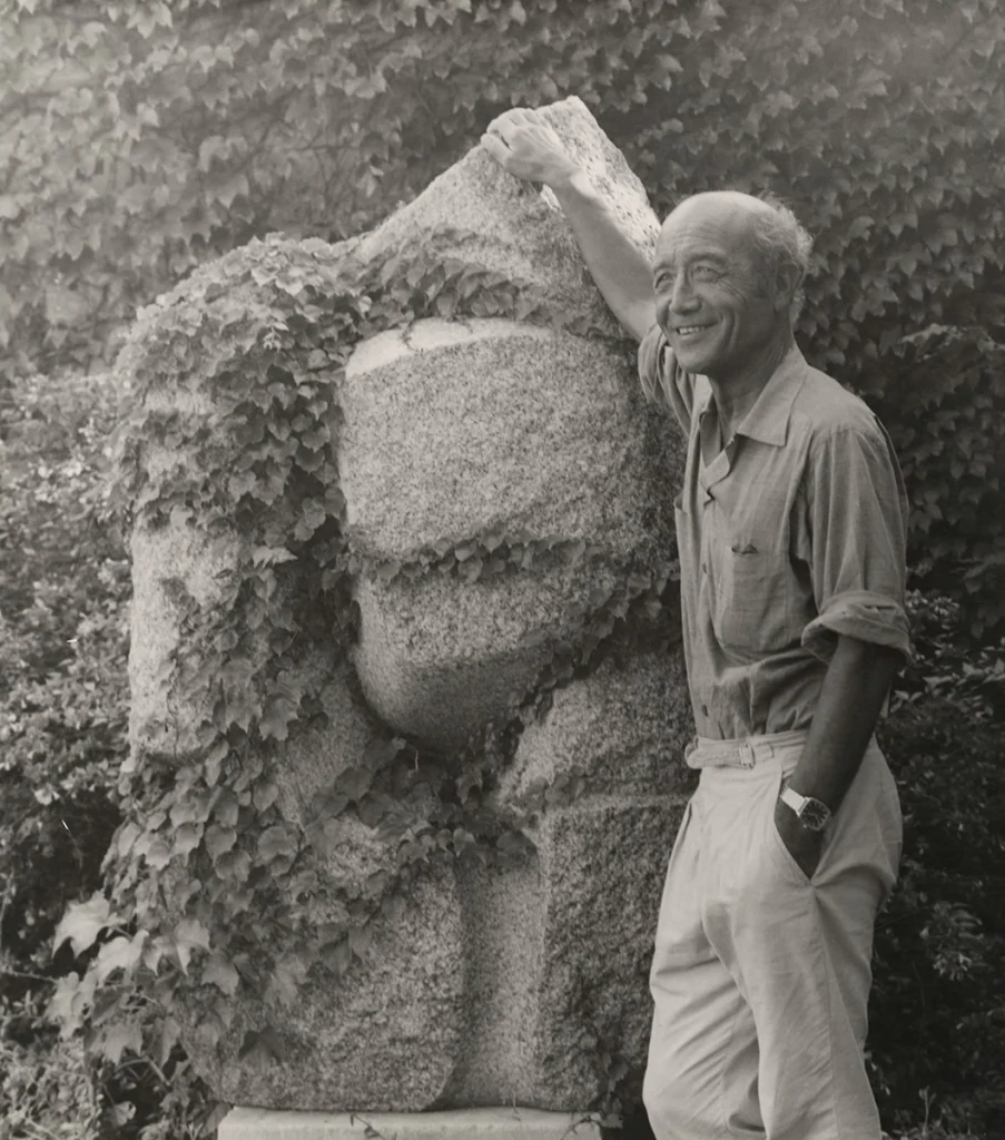 조각 작품 옆에 서서 미소짓는 이사무 노구치의 흑백 초상 사진 Black-and-white portrait of Isamu Noguchi standing beside a stone sculpture