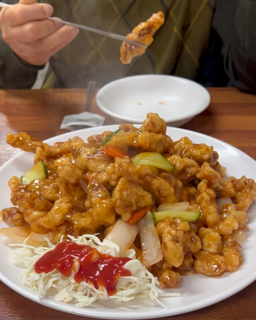 젓가락으로 목이버섯, 당근 등 소스가 코팅된 하림각 탕수육 고기 한 점을 집어 드는 모습. Picking up a piece of sweet and sour pork (tangsuyuk) coated in sauce with vegetables using chopsticks at Harimgak.