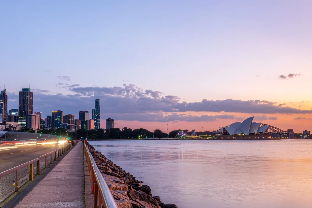 해질녘 시드니 오페라 하우스와 하버브리지, 도시 스카이라인이 보이는 풍경. The Sydney Opera House and city skyline at dusk, viewed from a pier.